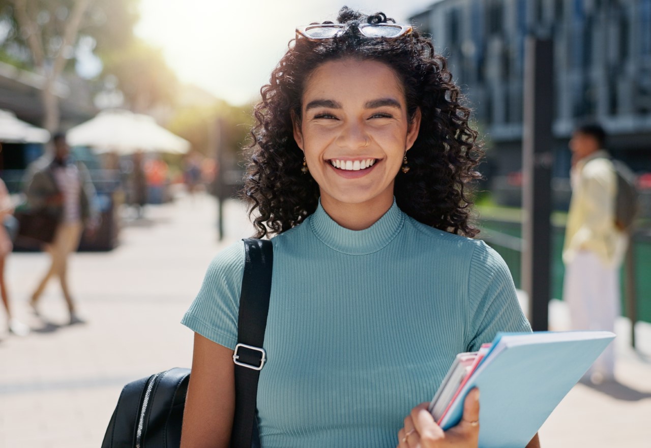 a young lady in front of a building smiling and holding a notebook, laptop and a tablet